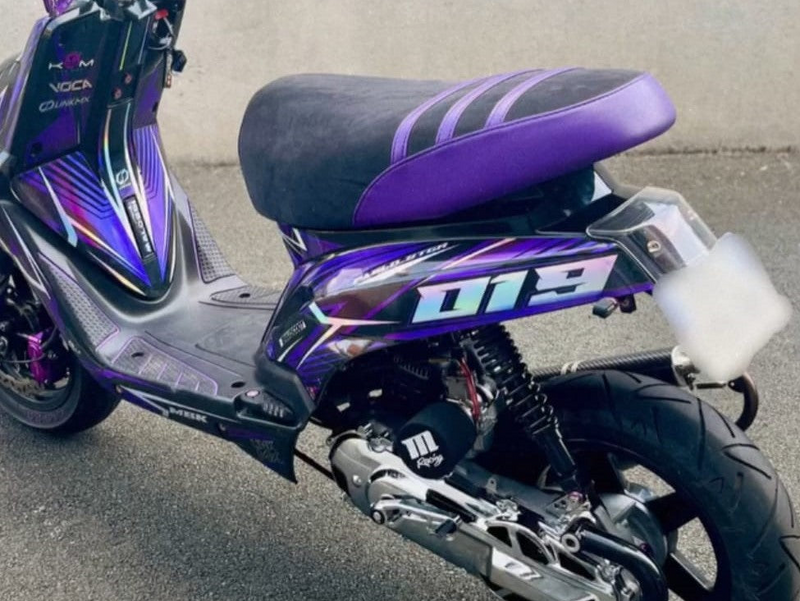 Un MBK Booster 50 violet à covering holographique, garé sur le bitume de l'atelier, équipé de la housse de selle BKS en daim noir et simili violet à bandes obliques