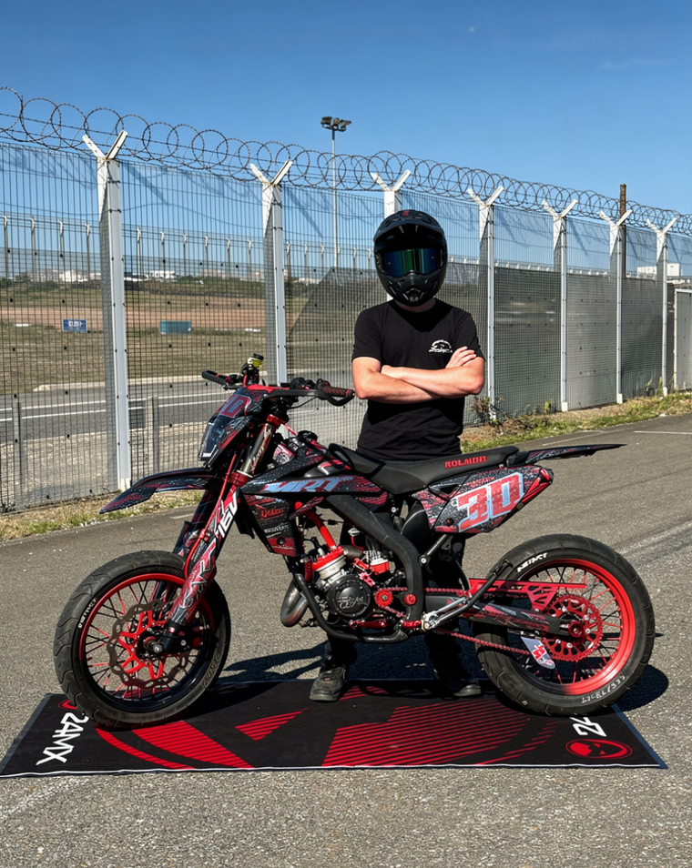 Moto rouge et noire numéro 30 sur un tapis de stand, pilote casqué derrière