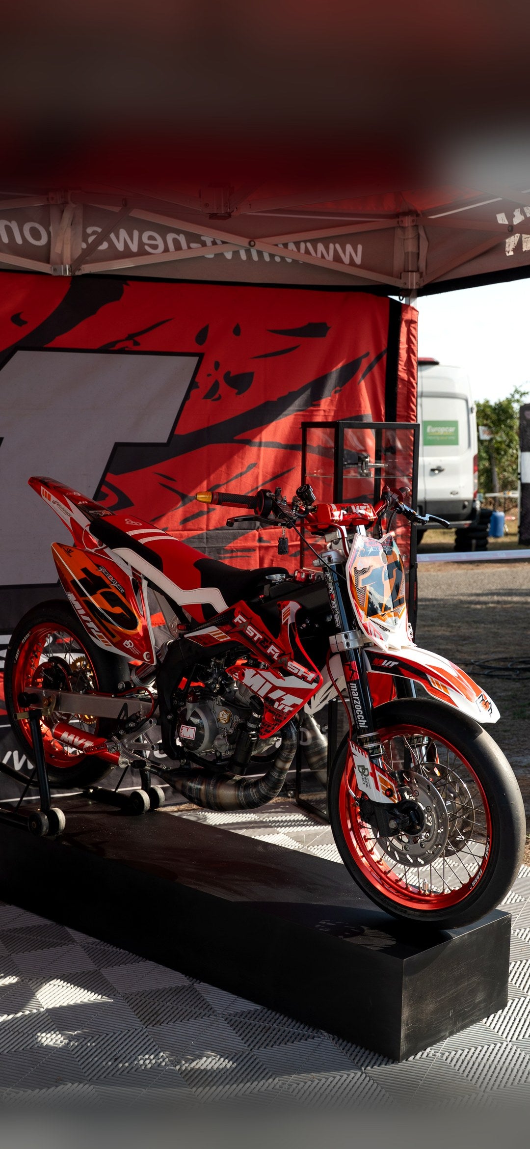 Moto 50cc rouge et blanche sur un podium, housse de selle BKS rouge à bandes blanches